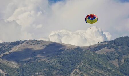 a multicolored parachute on a cable with passengers flies across the blue sky. Mountains in the...