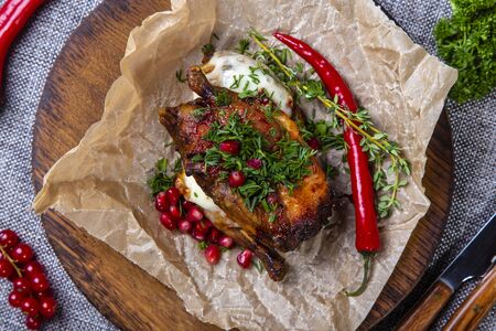 Baked chicken lies on a wooden tray wrapped in parchment paper.  The tray is on a gray canvas, next to a knife and fork, red pepper, currants.の写真素材
