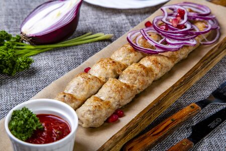 Lula-kebab of chicken with onions and hot red sauce. Served on a wooden tray. Background-grey linen. Around the cradle are spread onions, parsley, pomegranate and a knife and fork.の写真素材