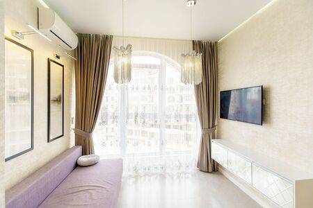 Kitchen room with a fresh renovation in beige tones. White kitchen set with kitchen appliances. semicircular window with curtains. TV, sofa and wicker furniture-table and two chairs.の写真素材