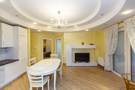 The hall is decorated in light colors. Oval table with chairs in the center. White leather sofa. Modern white kitchen. Round double ceiling with lighting and elegant chandelierの写真素材