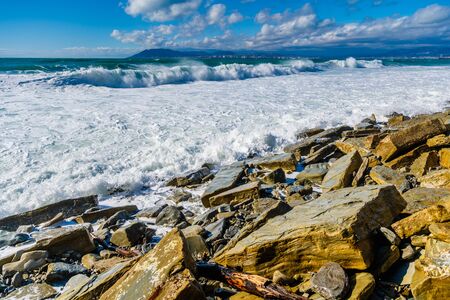 A storm surge in white sea foam hits the yellowish fragments of rocks on the beach. In the background Novorossiysk and mount Myshako.の写真素材