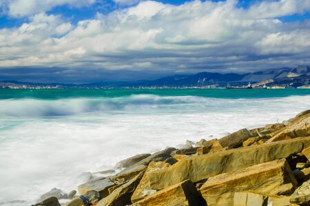 Storm waves at high speed are eroded and break in white foam on the beach, strewn with fragments of yellowish Sandstone rocks. In the background are mountains, a Bay and the port of Novorossiyskの写真素材