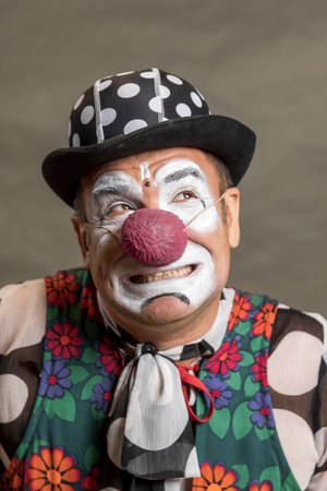 portrait of a funny clown in close-up. A clown in a black hat with white polka dots and a colorful vest makes faces. Photo on a gray backgroundの写真素材