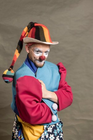 A clown with a white hat and red nose is holding a white jacket. The clown is wearing a yellow and...