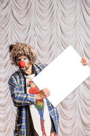 A funny clown holds an empty cardboard box on which you can paste text. A clown on the background of beige stage scenes.の写真素材