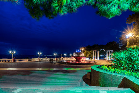 Gelendzhik resort embankment in the evening. There is a musical fountain in the center. Behind it is a colonnade. Balustrade along the sea line. The fountain is surrounded by a green bush. The walkway is lit up at night, creating a peaceful and serene atmosphereの写真素材