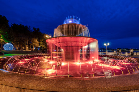 A large fountain with red water is lit up at night. The fountain is surrounded by benches and a few people are sitting on them. The scene has a peaceful and relaxing atmosphereの写真素材