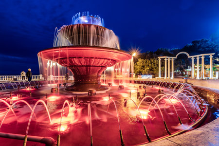 Gelendzhik resort, Black Sea, musical fountain. A large fountain with red water is lit up at night. The fountain is surrounded by benches and a few people are sitting on them. The scene has a peaceful and relaxing atmosphereの写真素材