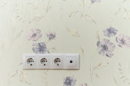 A white wall with a floral pattern and a white electrical outlet. The outlet is a three-pronged one
