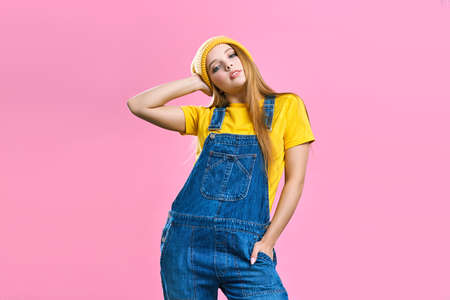 A pretty girl in denim overalls and a yellow hat on a pink background. Fashionista lady student. Bright trendy studio fashion image of sexy model, wearing neon bright color block clothes, casual vintage spring style, color pop, pink background, hipster girl posing and having fun.の写真素材