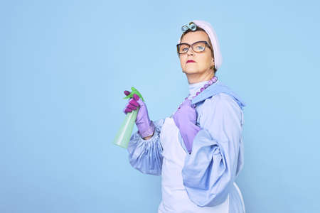 Portrait of old cleaning woman in apron with dust cleaning brush isolated on blue background. special uniform and professional equipment for cleaningの写真素材