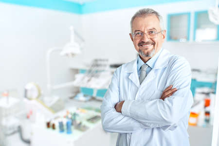 Portrait of glad smiling doctor in white uniform standing with crossed hands on blue dental clinic backgroundの写真素材