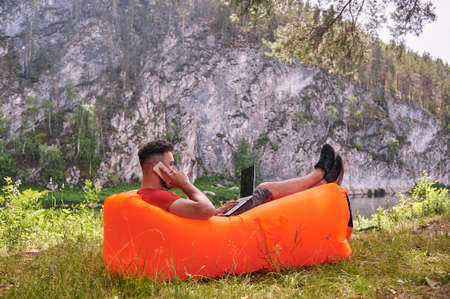 Bearded man is working and smiling with laptop at the park under the tree. Happy freelancer is sitting and using app or website on the grass. Work remotely in self-isolation modeの写真素材