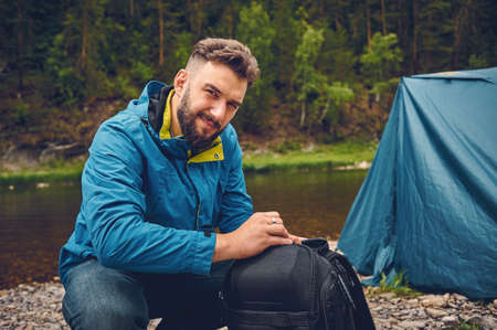 A young, bearded man with a backpack, camping, outdoors, against the backdrop of a tourist tent.