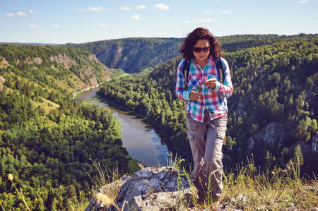 A girl with a backpack stands on a rock on a background of a valley and holds flowers in her hand.の写真素材
