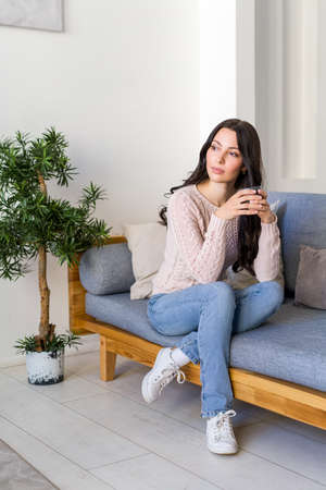 Young woman drinking tea and dreaming sitting on the couch in the bright apartment.