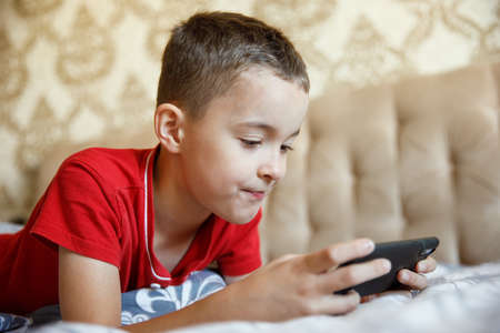 The boy is playing games on his smartphone, lying on the couch at home.