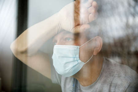 Man with a face mask sitting in a home quarantine and looking through the window.の写真素材