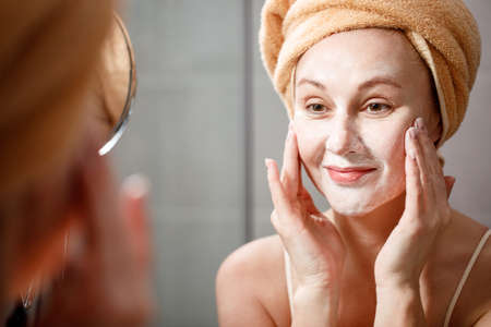 A woman puts a cleansing mask on her face in the bathroom in front of the mirror. Skin care....