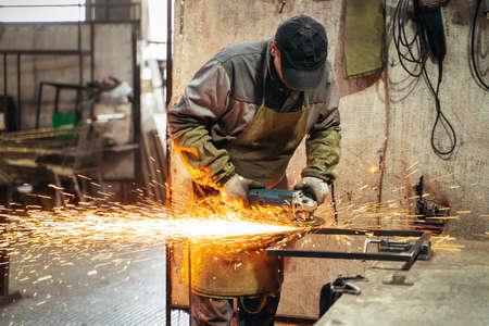 Locksmith in special clothes and goggles works in production. Metal processing with angle grinder....