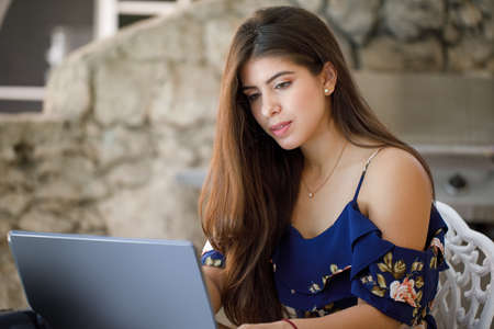 Millennial latin girl sit at desk in outdoor on laptop making notes. Concentrated young woman work on computer. Take online course or training. Education concept.の写真素材