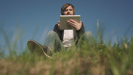Bottom View of Handsome Hipster Using Tablet PC While Sitting On The Grass In The Park On A Sunny Day, Outdoor, 4K, UHDの写真素材