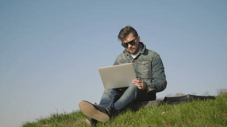 Young Hipster Man In Sunglasses Using Laptop Outdoors Sitting On The Grass In Park, 4K, UHDの写真素材