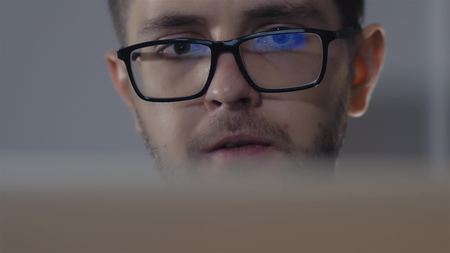 Man in black rim glasses using his tablet computer in dark room. Screen reflection in the lens