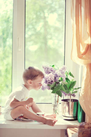The child sits on a window sill at a window near to a lilac and smells aroma of flowersの写真素材
