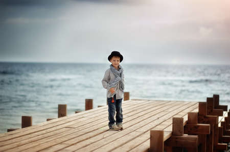 Stylish boy in a hat standing on a wooden bridge on the background of the seaの写真素材