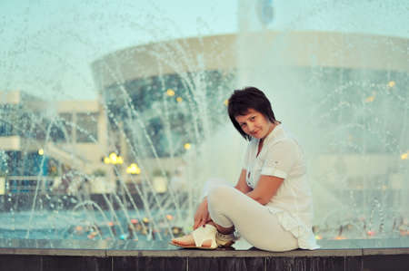 against splashing fountain sits a woman in a white suitの写真素材