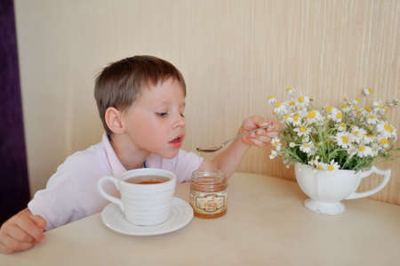 little boy drinks tea and eats with a spoon and honey on the table a bouquet of daisiesの写真素材