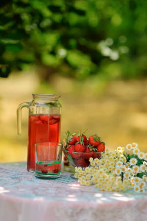 on a white table is a jar of strawberry compote and glasses and a bouquet of daisies and a bowl of...
