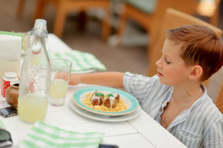 at a table in a cafe boy in white shirt eats noodles with meatballsの写真素材