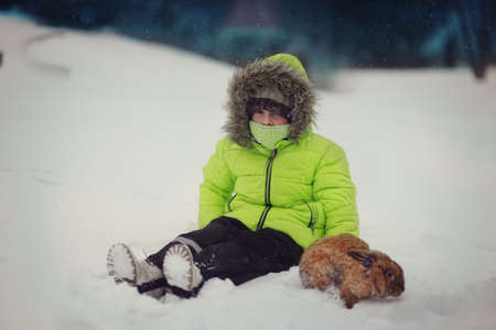 cute boy in green jacket sitting in the snow with a rabbitの写真素材