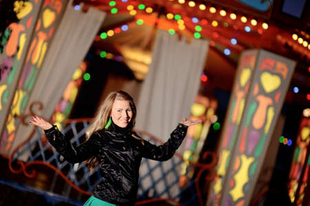 blonde girl with long hair in black jacket in the evening on the town stands in front of a colorful carouselの写真素材