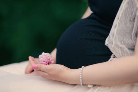 Pregnant woman in black dress hugs belly and holding a flower