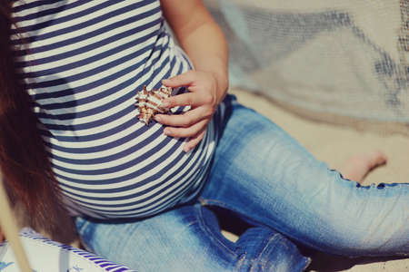 Pregnant woman in jeans and a vest hugs belly and holding a seashellの写真素材