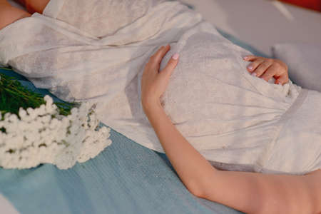 on a blue bedspread is a pregnant woman in a white dress with a bouquet of flowersの写真素材