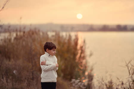 against the sunset over the river near the cattails stands cute boy in white sweaterの写真素材