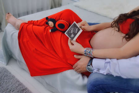 belly of a pregnant woman in a red dress and in the hands holding ultrasound photoの写真素材