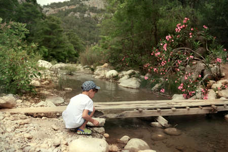 in the woods near a mountain river on the bridge sits a little boy in a capの写真素材