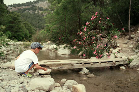 in the woods near a mountain river on the bridge sits a little boy in a capの写真素材