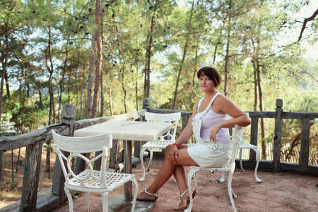 in a pine forest sits a woman in a hat on a chair at the table of an old fenceの写真素材
