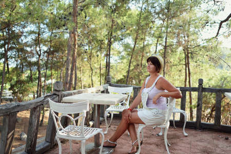 in a pine forest sits a woman in a hat on a chair at the table of an old fenceの写真素材