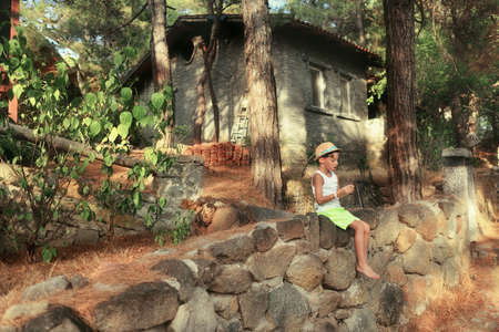in the woods near the house sits a boy in a hat on a stone fenceの写真素材