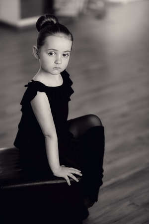 little ballerina in a black dress in pantyhose sitting on a black square in the hallの写真素材