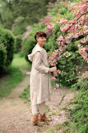 Woman with stylish hairstyle and light raincoat in the lush garden near the cherryの写真素材
