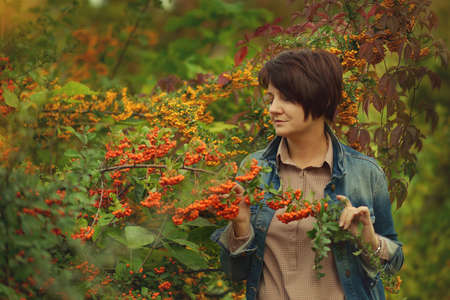 woman in a denim jacket against a background of large green bush with red rowanの写真素材